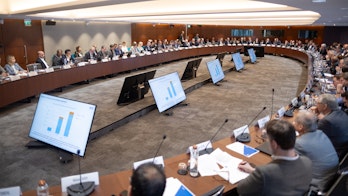 Image shows a large conference room with rounded tables across the edges with people sitting in the chairs