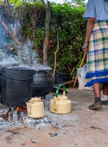 Photo depicts a woman cooking using four black cauldrons over an ashy and wood filled fire