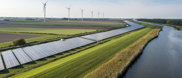 Photo shows a green landscape with a row of modern wind mills and solar panels to produce clean energy. A canal of water flows on the right side of the photo into the horizon,