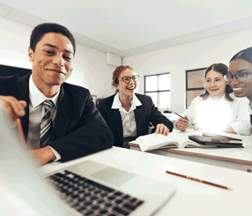 Teenage boy and girl students, wearing school uniforms, smiling together while sitting around a computer at a desk in a classroom.