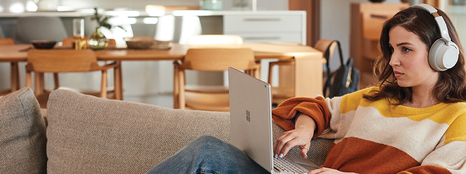 A woman uses a Surface laptop and Surface Headphones while lounging on a couch at home.