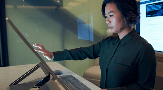 ઝાંખો પ્રકાશ ધરાવતી ઑફિસમાં Microsoft Surface Studio પર સ્ક્રોલિંગ અથવા કાર્ય કરતી ડાર્ક શર્ટ પહેરેલી સ્ત્રીની સાઇડ પ્રોફાઇલ.