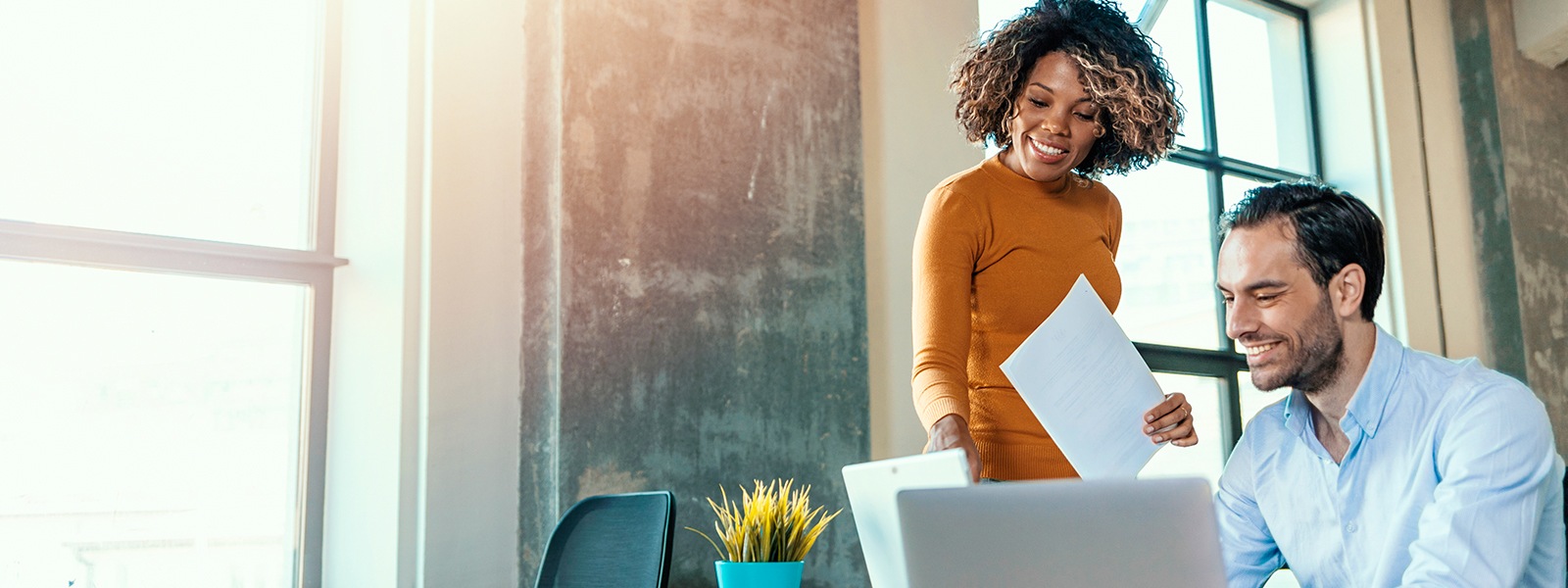 Woman standing in an office looks at a laptop screen with a seated male colleague.