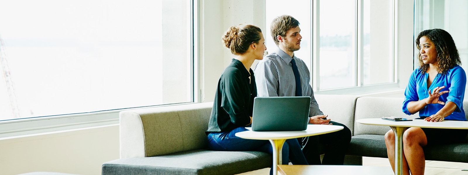 A businesswoman leads a meeting with two colleagues in a sunny office.