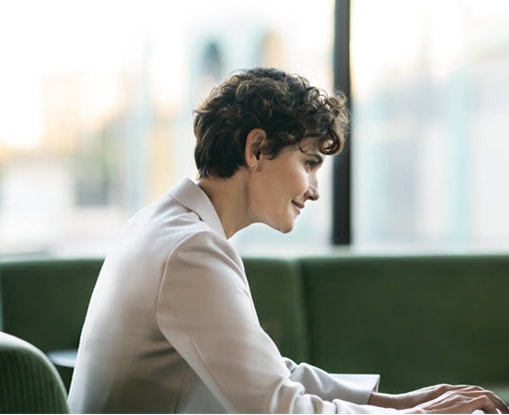 A woman types on her laptop while sitting outside in a natural area.