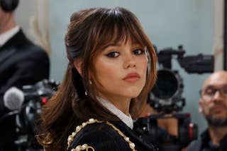 Jenna Ortega on a red carpet wearing a black, intricately designed outfit adorned with pearls and gold chains, with people and cameras in the background