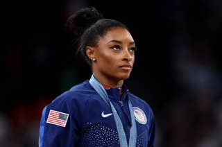 Simone Biles stands wearing a Team USA jacket, a medal around her neck, and an American flag patch on her sleeve