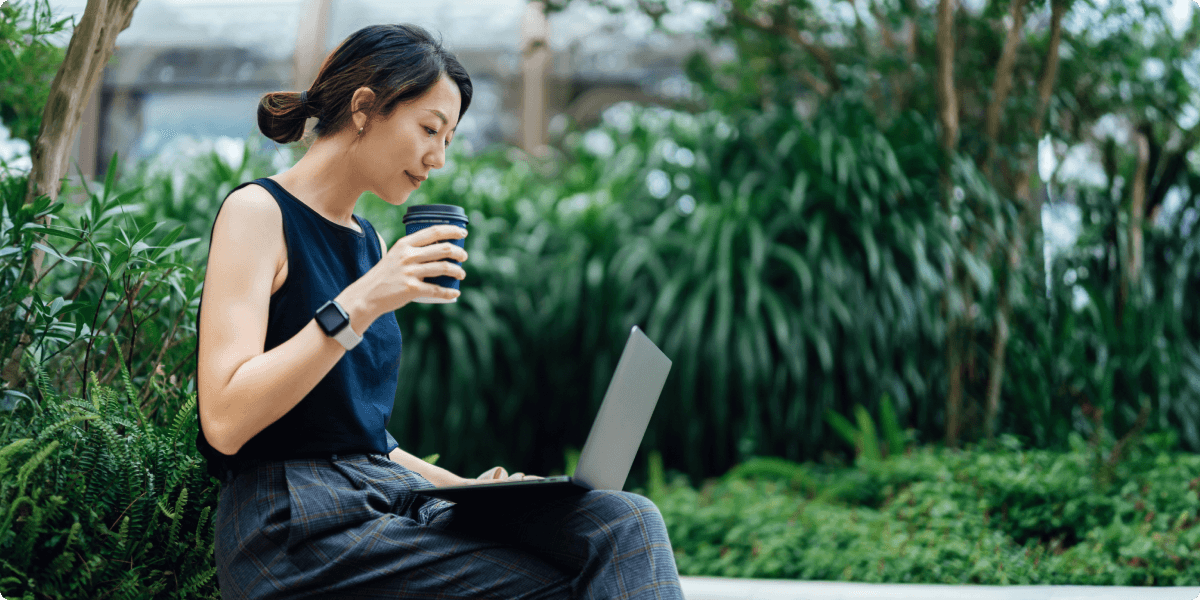 En kvinde på sin bærbare computer udenfor med en kop kaffe. Hun er omkring frodige grønne planter.