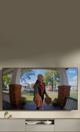 A TV in a living room showing a delivery person standing on a stoop waving to the doorbell camera.