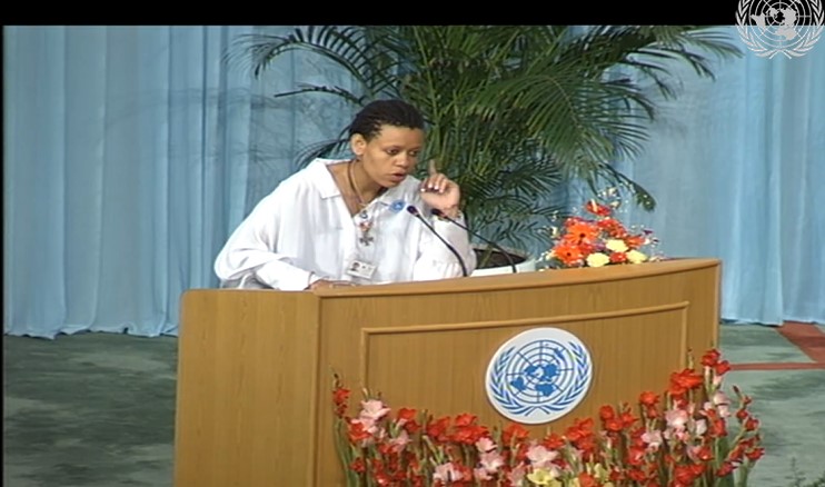 Palesa Beverly Ditsie gives a speech, leaning over a podium with her index finger pointed up. She stands behind a podium with the UN logo.