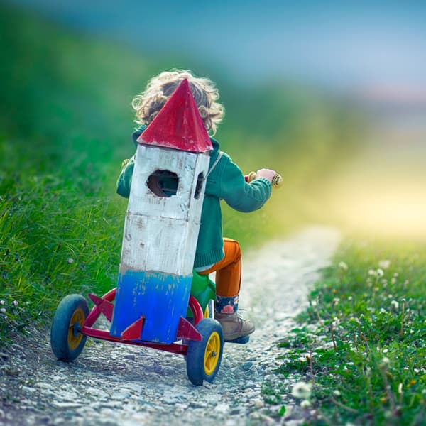 A boy riding his tricycle with a cardboard rocket strapped to his back