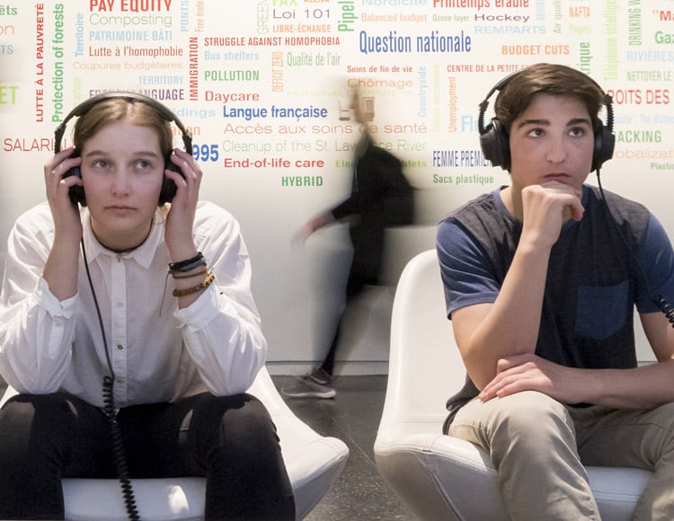 Jeunes adultes assis dans des fauteuils blanc écoutant et regardant une vidéo dans une exposition.