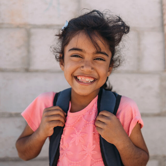 A child is holding a 	backpack and smiling. 