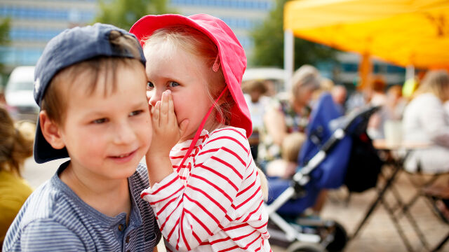 Kaksi lasta kuiskuttelee toisilleen torilla. 