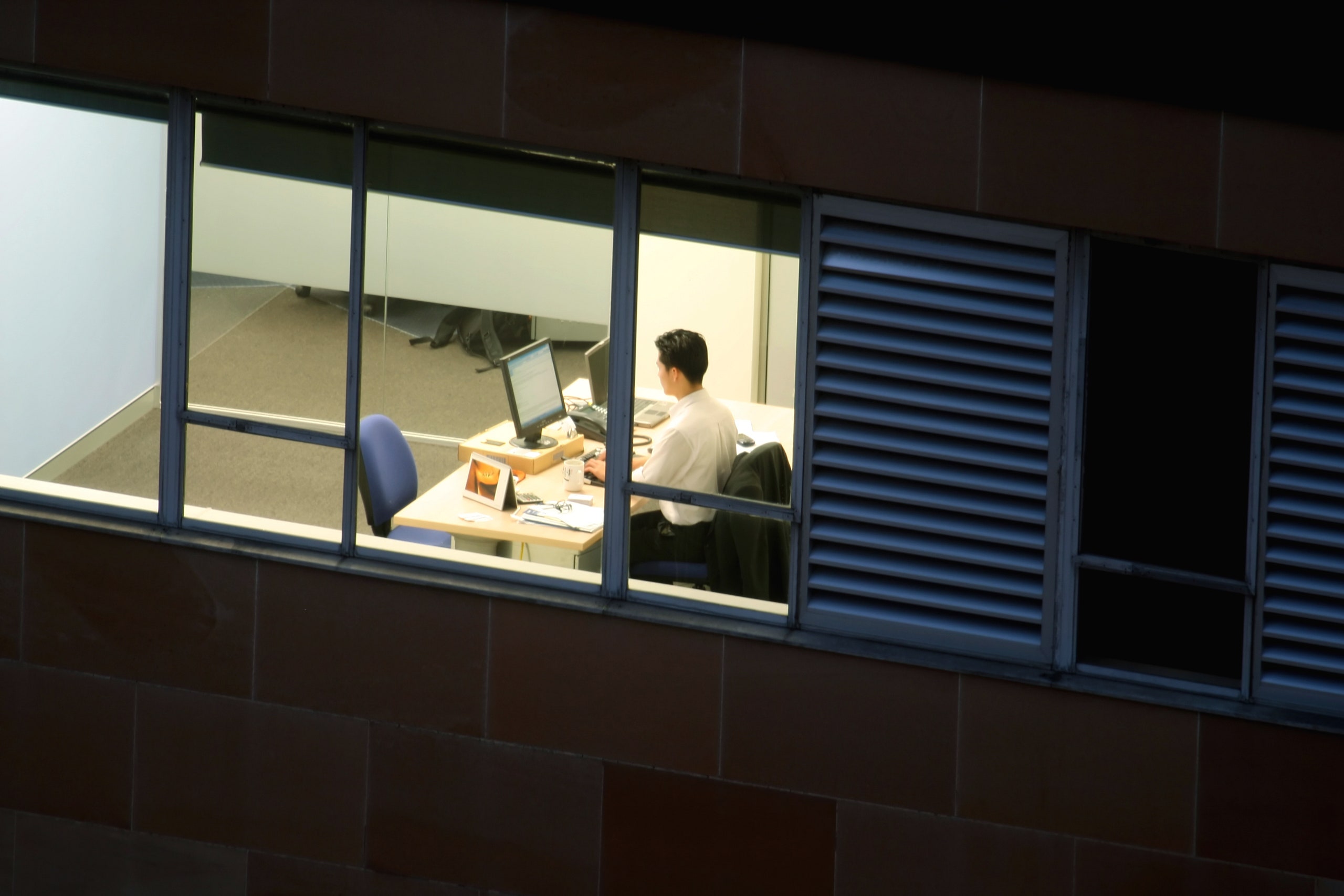 A person sits alone at his computer in a brightly lit office at night.