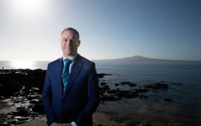 Climate Change minister Simon Watts, wearing a blue suit, stands in bright sunlight near a rocky shore, with Rangitoto Island in the background.