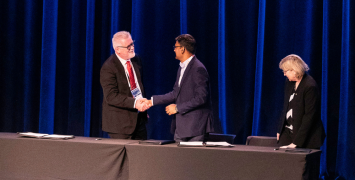 Three people standing by a desk on stage. Two are shaking hands. 