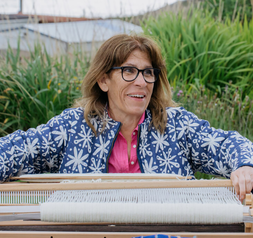A smiling woman in a blue-and-white floral jacket works at a traditional loom that is flat on a table.