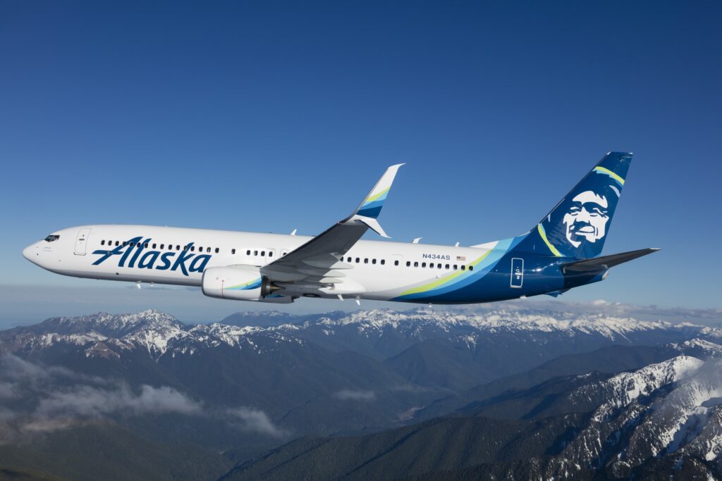 An Alaska Airlines aircraft flies over a snow-capped mountain range.