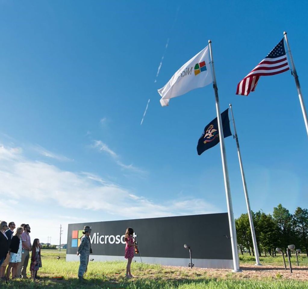 A group of people gathers below flags flying above the Microsoft campus
