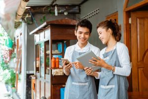 photograph of a man and woman wearing aprons, looking at a tablet with a wooden food stall behind them