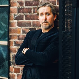 Josh Boldt wearing a black corduroy blazer with his arms crossed in front. His back is leaned against a brick wall.