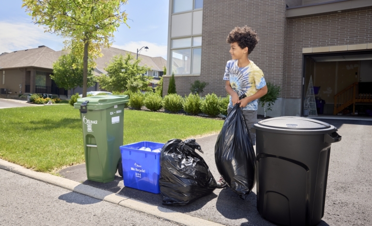 Un enfant tenant un sac noir à côté d'un bac de recyclage, d'un bac vert et d'un contenant de déchets.