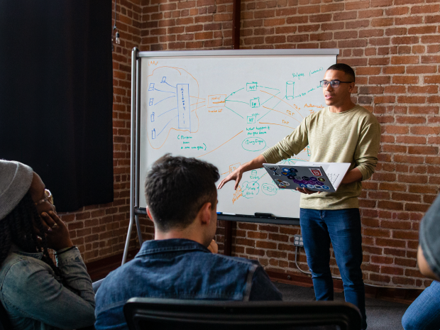 Man presents in a meeting from a laptop and whiteboard to his coworkers