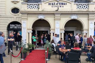 A red carpet leads into the entrance of the cinema. People are gathered outside, chatting in groups.