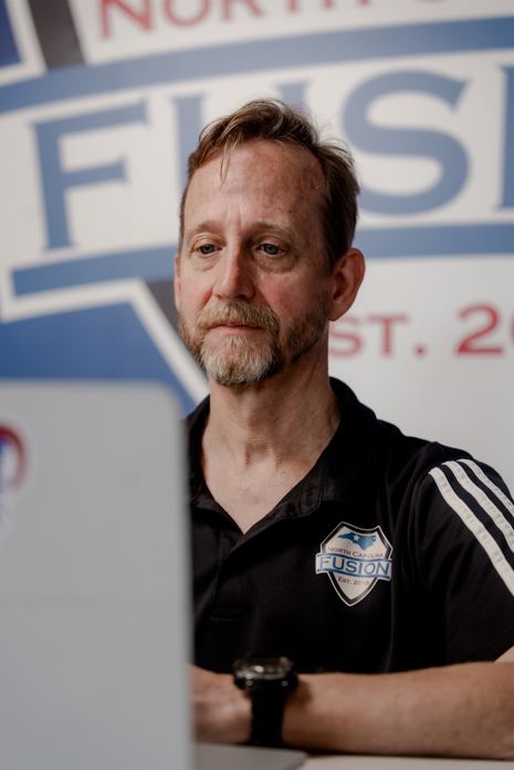 Chris Barnhart is looking at his laptop screen while while sitting at a table in front of an NC Fusion sign.