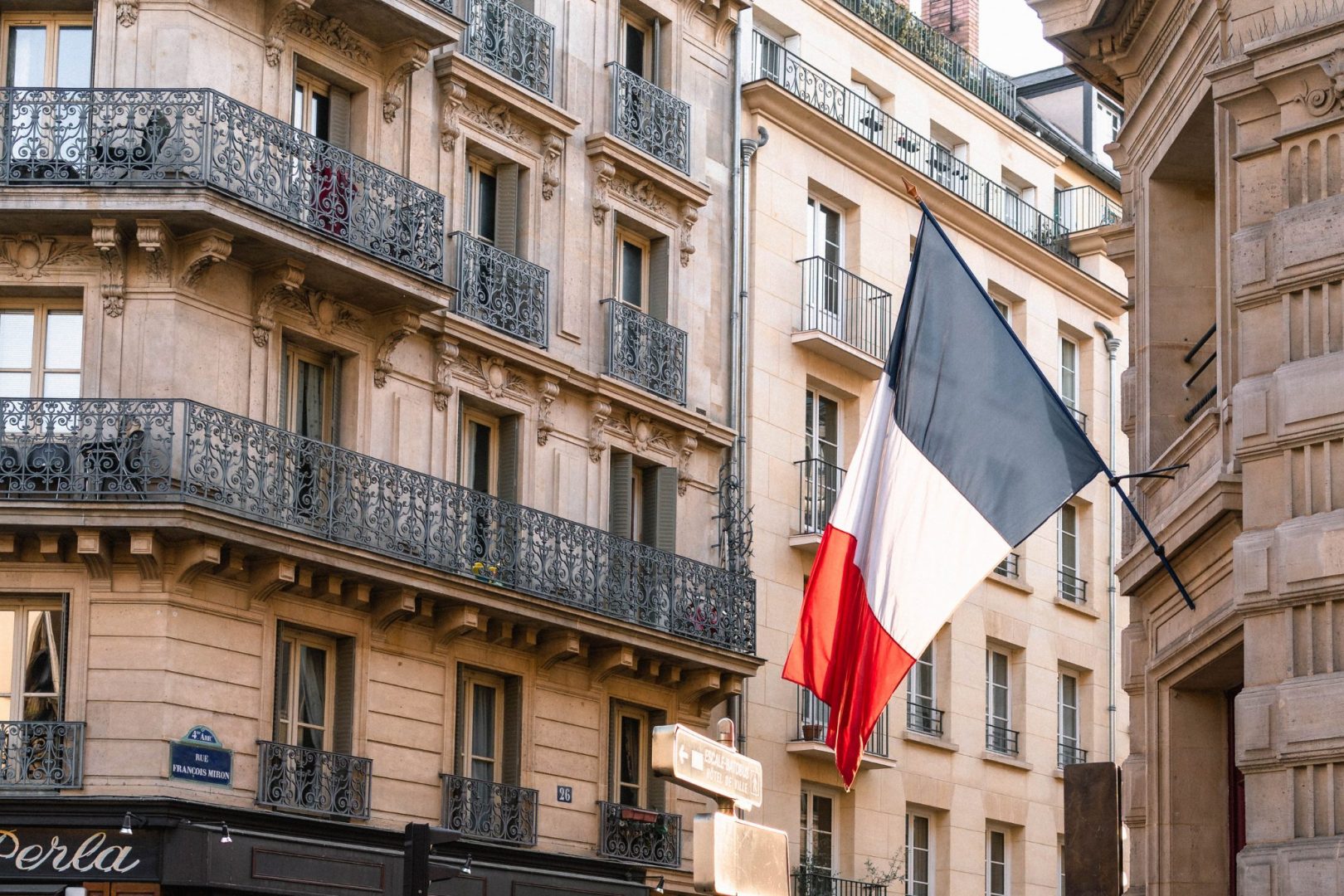 French flag hanging from a pole on a traditional Parisian building with ornate balconies and adjacent street signs.