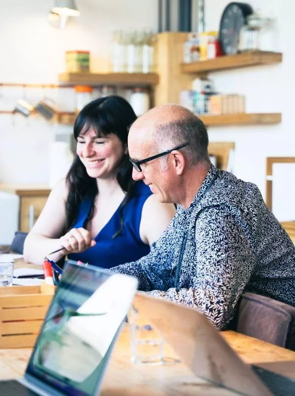 A designer is wearing a bold patterned shirt and glasses. He is sitting with a woman with long dark hair. Both are smiling and looking at a screen on a desk. Two more laptop screens can be seen in the foreground while in the background is a blurred out studio office kitchen.