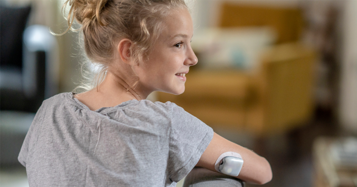 young blonde girl wearing a gray tee shirt is sitting with her arm visible. She's turned to the back right and we can see her diabetes monitor on the back of her right arm