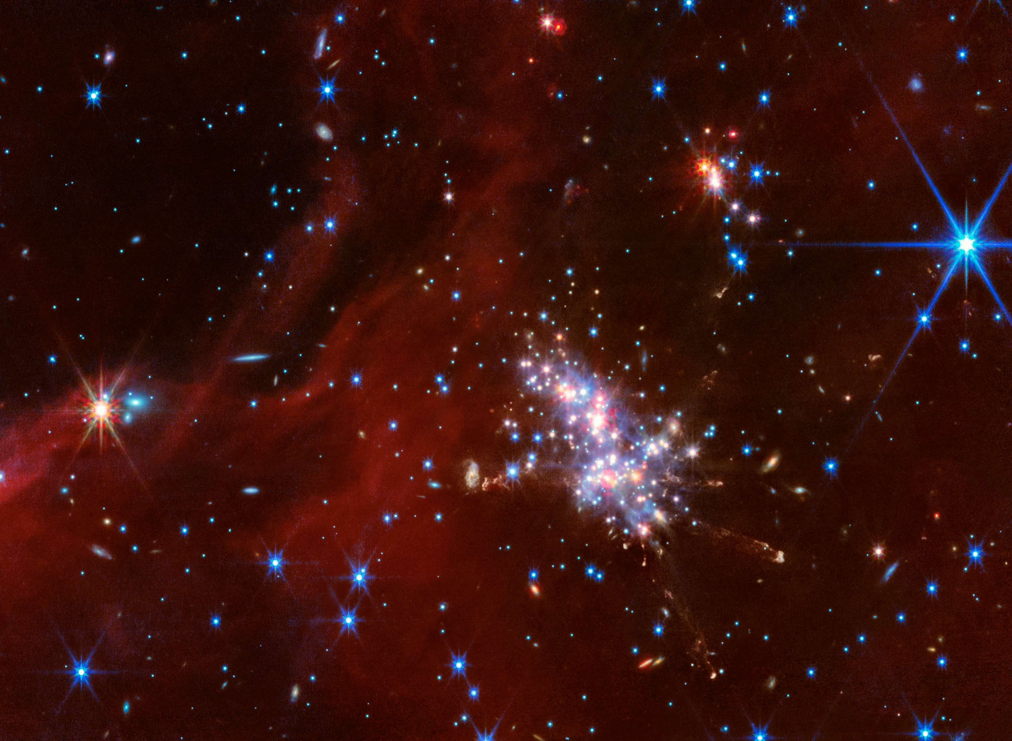 At center right is a compact star cluster composed of luminous red, blue, and white points of light. Faint jets with clumpy, diffuse material extend in various directions from the bright cluster. Above and to the right is a smaller cluster of stars. Translucent red wisps of material stretch across the scene, though there are patches and a noticeable gap in the top left corner that reveal the black background of space. Background galaxies are scattered across this swath of space, appearing as small blue-white and orange-white dots or fuzzy, thin disks. There are two noticeably larger points, foreground stars, with diffraction spikes: an orange-white point on the left, and a blue-white point in the top right.