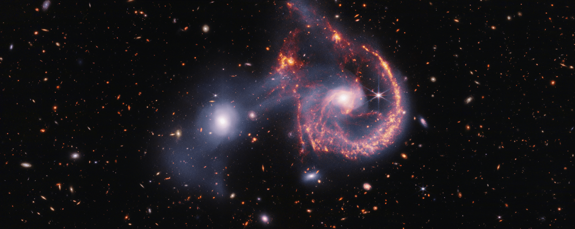 A pair of interacting galaxies. The larger of the two galaxies is slightly right of center, and composed of a hazy, bright, white center and a ring of gaseous filaments, which are different shades of red and orange. Toward the bottom left and bottom right of the ring are filaments of gas spiraling inward toward the core. At the top left of the ring is a noticeable gap, bordered by two large, orange pockets of dust and gas. The smaller galaxy to its left is made of hazy white gas and dust, which becomes more diffuse farther away from its center. To this galaxy’s bottom left, there is a smaller, more diffuse gas cloud that wafts outward toward the edges. Many red, orange, and white galaxies are spread throughout, with some hazier in composition and others having more defined spiral patterns.