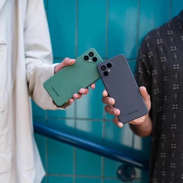 Two hands presenting Fairphones with distinctive cases, one in dark grey and another in speckled green, against a vibrant blue tiled background, emphasizing the stylish and sustainable design of these eco-friendly smartphones.