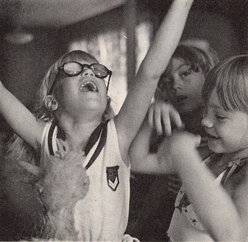 Three young white girls are playing. One girl with glasses has her arms and head raised joyously up towards the ceiling. A second girl is smiling and is in the middle of extending her arms up as well. A stuffed animal is visible in the foreground.