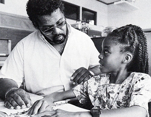 A young Black girl and her father sit side by side as the girl reads braille text on the desk in front of her.