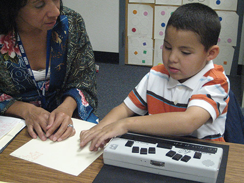 A young boy who is blind and a female teacher sit behind a desk in a classroom. A braille writer is infront of the student. He is reading text in braille that is on the table beside the braille writer. His teacher looks on.