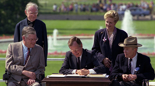 President George H. W. Bush sits at a desk on the South Lawn of the White House. Seated in wheelchairs are Evan Kemp to his right and Justin Dart to his left. Standing behind them are Reverend Harold Wilke and Swift Parrino.