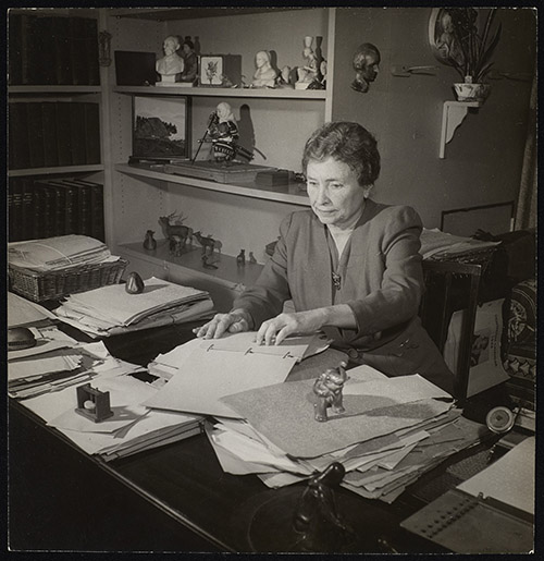 Helen Keller sits at her desk in her home office. She is reading a document in braille. Papers and objects cover the table and books and objects are visible on shelves behind her.