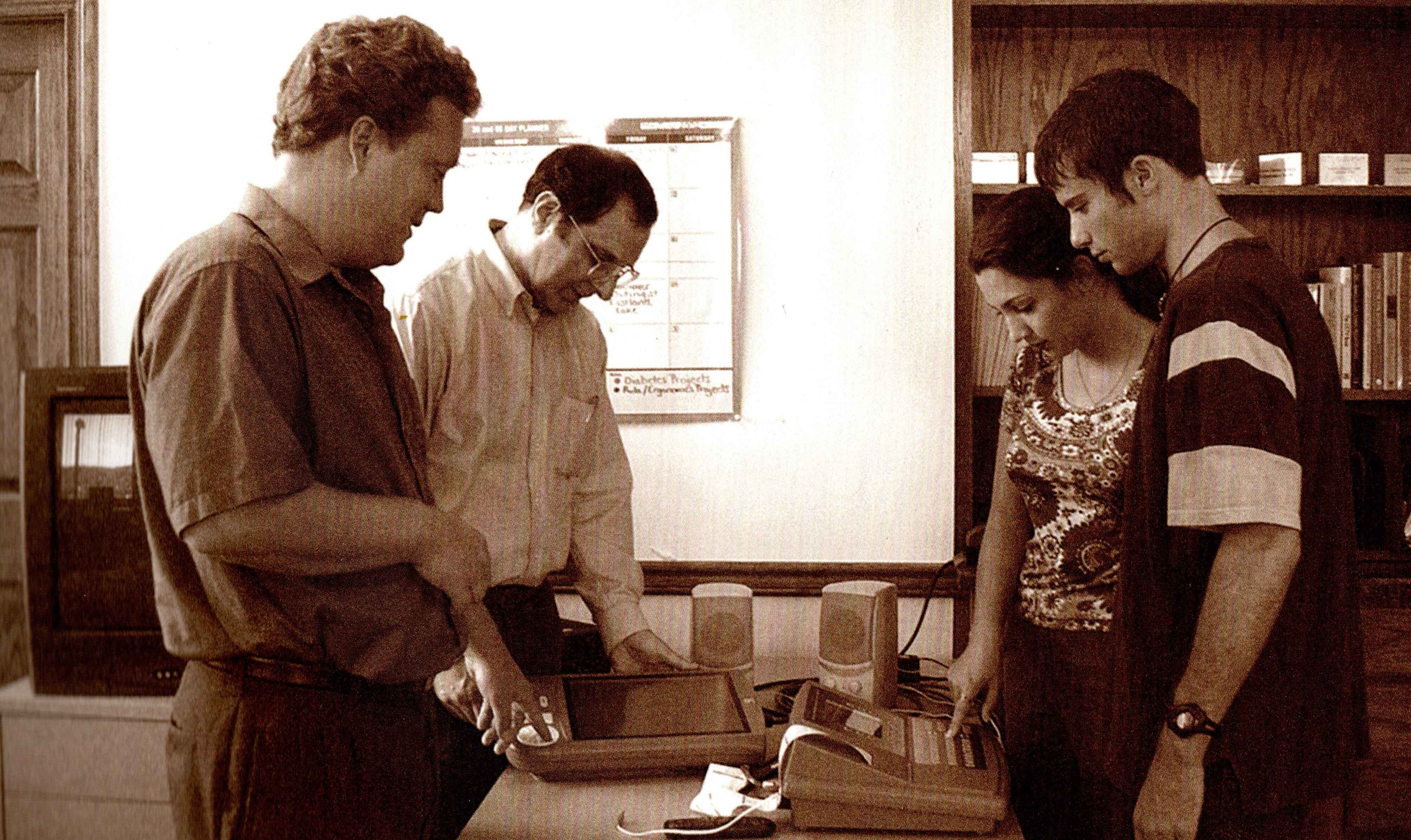 AFB Tech staff Darren Burton and Mark Uslan, both white, evaluate accessible voting machines that are on a table. Two interns, a woman and a man who are both white, assist them.