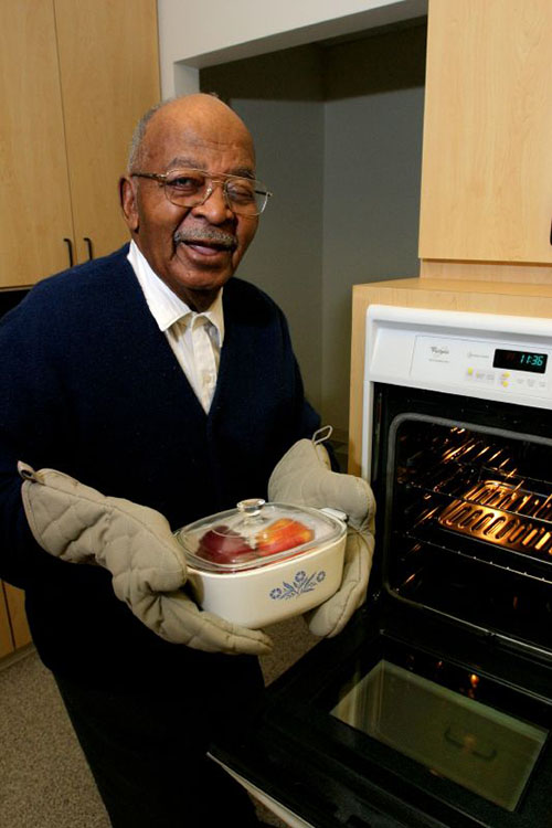 An elderly Black man in a kitchen smiles at the camera as he holds up a square white casserole dish with a clear glass lid in his gloved hands. He stands infront of the open door of an oven.