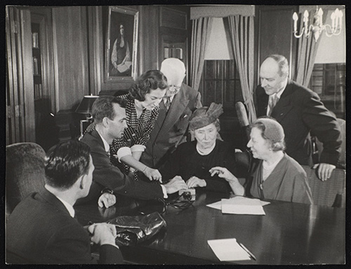 In a wood-paneled room, several white people surround Helen Keller, who sits at a table with her hands on a small device.