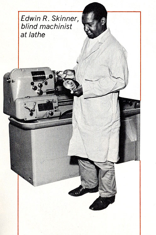 Seen in a laboratory coat, a blind Black man stands in front of a large factory lathe and holds a piece of equipment.