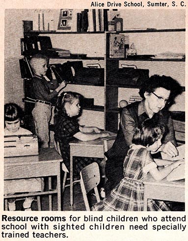 Four young, white school children who are blind study in a resource room. Three girls sit at individual desks. One uses a braille writer, while the other two read braille texts. A white female teacher sits next to one of the girls and helps her read. In the background, a boy selects books from bookshelves.