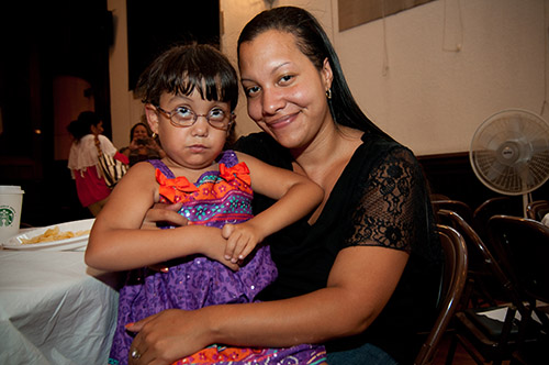 A young Brown girl wearing a purple and orange sleeveless dress with straps sits on a woman's lap. The child wears round glasses and is looking up. The woman smiles at the camera.