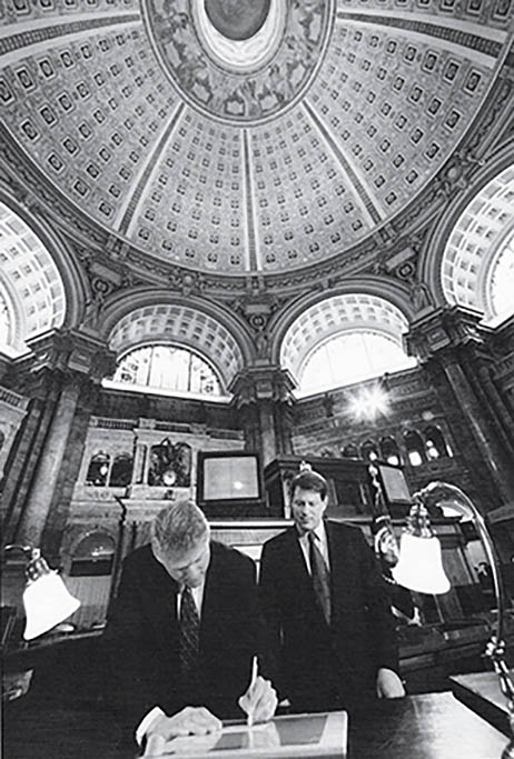 President Clinton, standing, signs the Telecommunications Act in a rotunda with arched windows and a beautifully decorated ceiling. Vice President Al Gore stands behind the President.