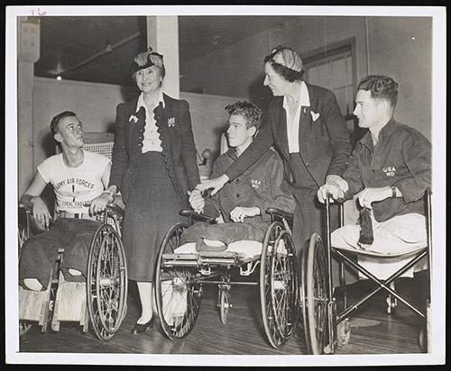 Three white men whose legs have been amputated, with Helen Keller and Polly Thomson. Keller, smiling, touches the hand of the man to her right and holds the hand of the man to her left. The veterans wear military hospital clothing and are using wheelchairs.