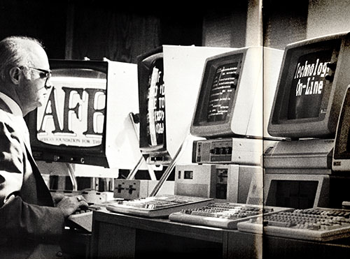 A white man wearing glasses stands at a computer monitor displaying magnified text. Additional monitors and computers are to the left and right of him, displaying text of various sizes, including a large AFB logo.